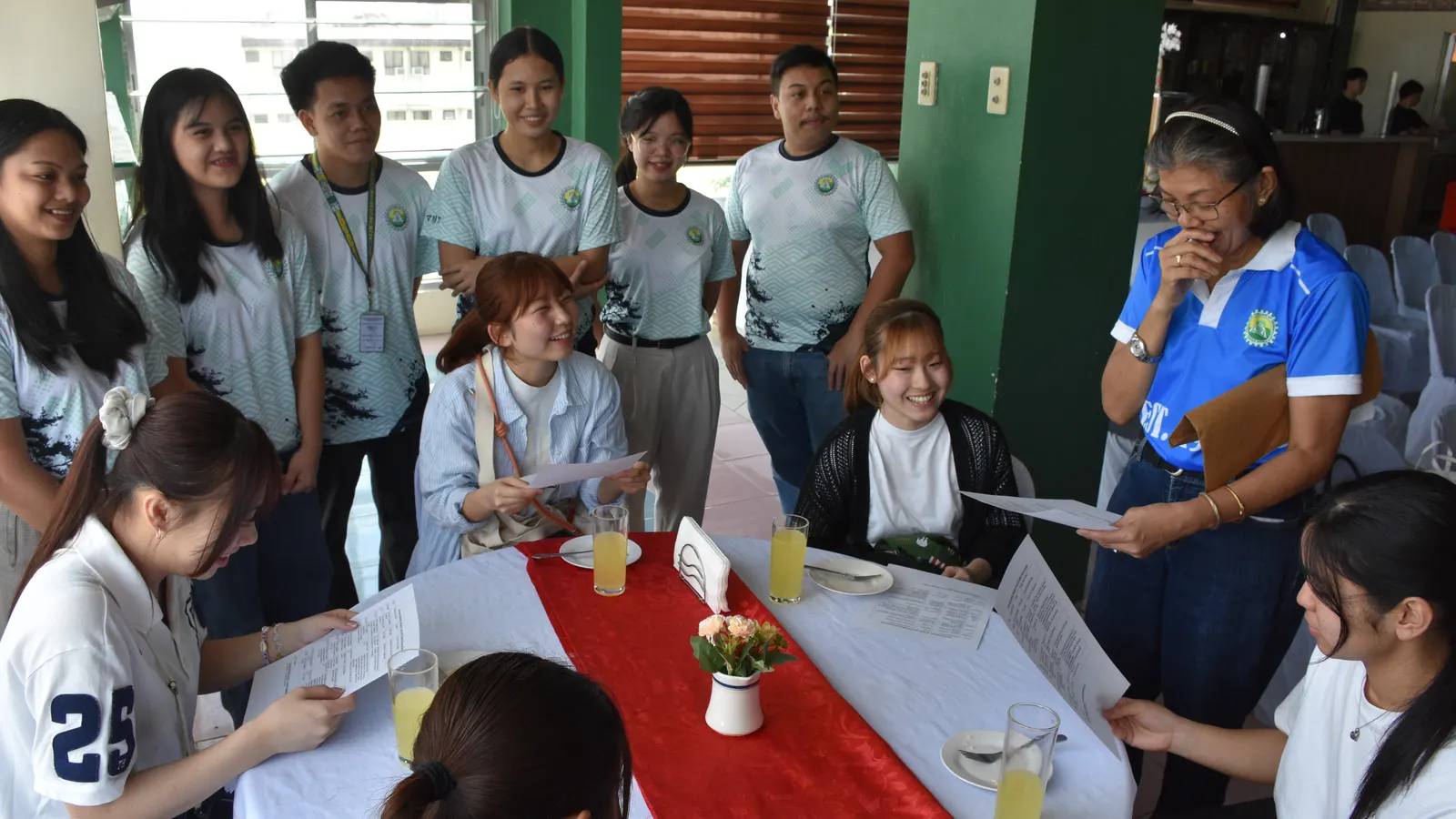 A group of students and a teacher gathered closely around a table indoors, reviewing papers together and smiling