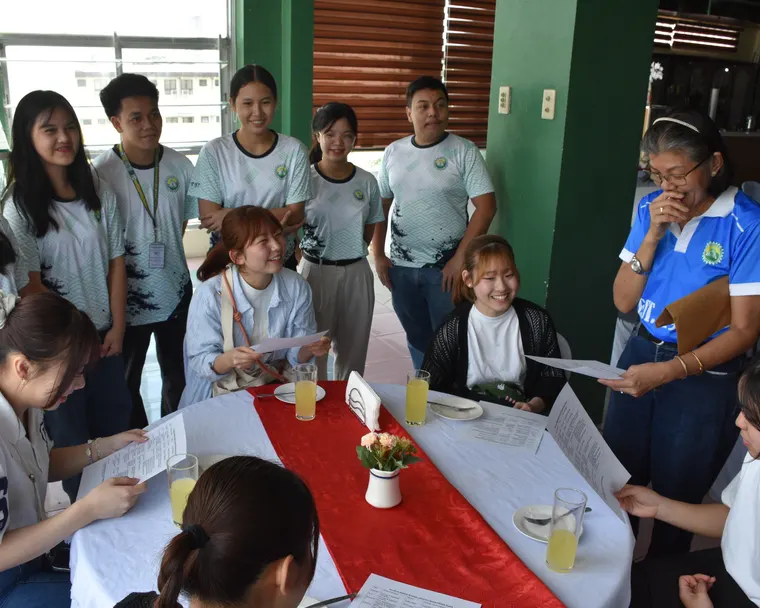 A group of students and a teacher gathered closely around a table indoors, reviewing papers together and smiling