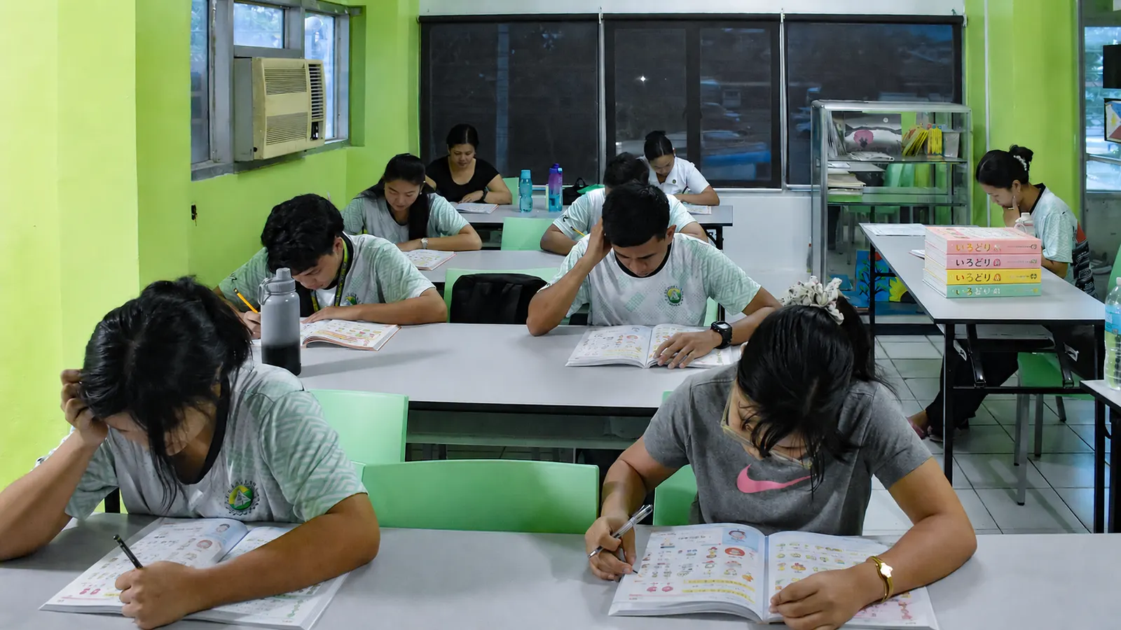 Students at classroom desks writing in Irodori workbooks, with stacked copies of the Irodori textbook series on a table nearby